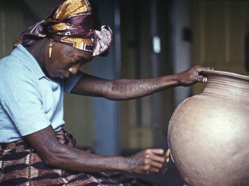 Photograph of ceramics artist Ladi Kwali. She is holding a large pot in one hand while inscribing on it with the other.