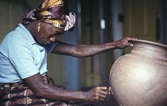 Photograph of ceramics artist Ladi Kwali. She is holding a large pot in one hand while inscribing on it with the other.