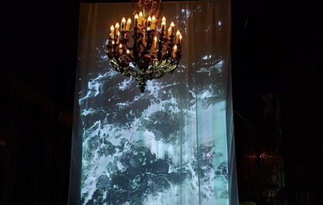 Image shows an instillation where a projected image of water from the river Thames has been put onto a piece of hanging fabric. In front is a gold chandelier.