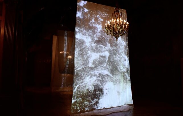 Image shows a large instillation, a video of the water from the River Thames has been projected onto a large piece of fabric. At the top of the image is a gold chandelier.