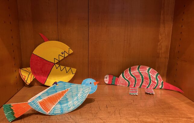 Image shows colourful paper plate animals on a wooden shelf.